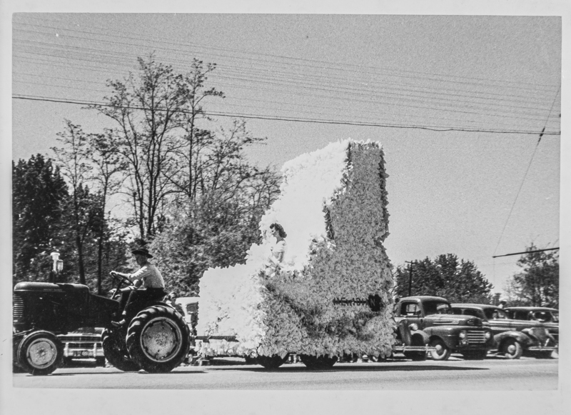 A float shaped as the state of Idaho. Riding the float is a young woman, perhaps a beauty queen. The float is being pulled by a tractor.