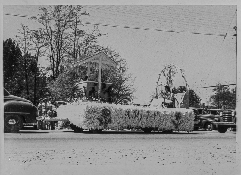 A young lady riding a float with "Amity" lettering. She is holding a parasol. The float is pulled by an automobile.