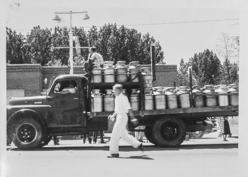 A milk hauler drives his truck filled with milk cans. A boy rides atop the cans while another man walks beside the truck.