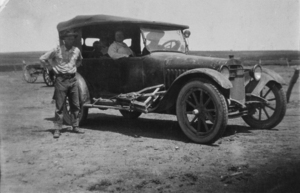 Car in the middle of a field with five people. One outside the car, two up front, two in back.
