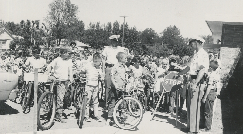 Police Chief Gene Hiner leads bicycle training course at Meridian grade school. Upon completing course, children issued bicycle safety certificates signed by Hiner and school principal Obed Dahl