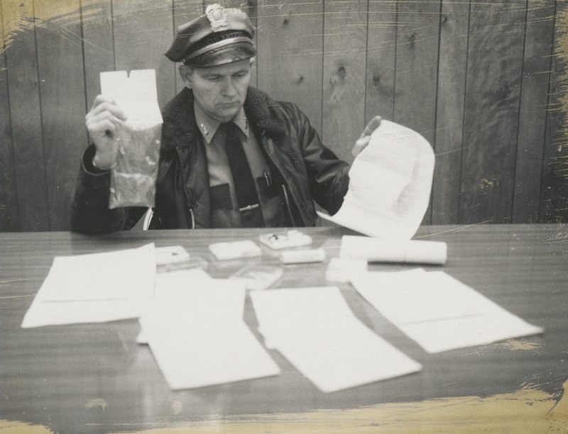 Circa 1970, Gene Hiner examines evidence in a drug case (bag in right hand contains marijuana)