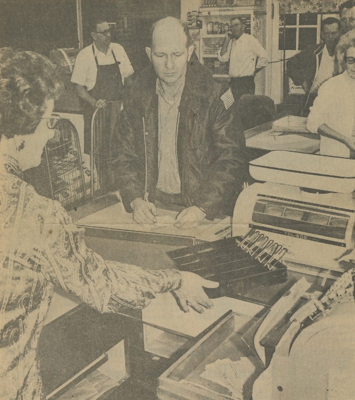 Davis Food Store clerk, Jessie Jackson, shows Police Chief Hiner that the robbery suspects did not get a stack of five dollar bills under the cash tray, although they took the other cash. George Davis, store owner, is on the phone in the background.