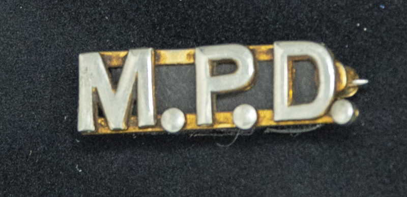 Gene Hiner MPD collar pin, the first part of his MPD collar pin set. The matching collar pin says "Chief." The MPD and Chief pins would be pined, one on each side of the shirt collar. Hiner would have worn this between 1963-1973. Part of the Gene Hiner Police Career Display presented to the MPD by Gene Hiner's family. 