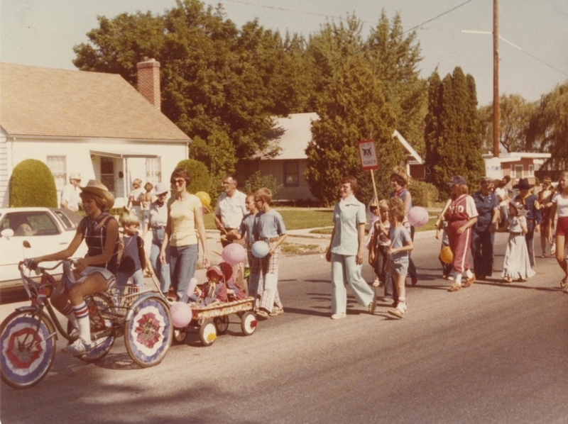 Meridian's 75th Birthday Parade, July 27, 1978. Laura Rabehl can be seen on the three-wheel bike with her twin daughters in the wagon; Eleanor Arndt is walking in the jeans and cowboy hat; Nancy Spear is in the light blue pantsuit; Pat Rabehl is in the straw hat; Shelly Olsen is in the baseball uniform; and Fern Wilson is in the dark blue pantsuit. A child can be seen holding up a sign with a picture of Figaro, the Meridian Library Cat. 