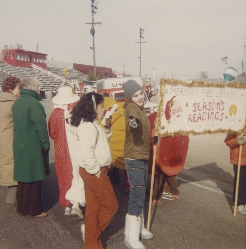 Meridian Library parade walkers prepare for Holiday Parade, holding sign that says "Season's Readings."