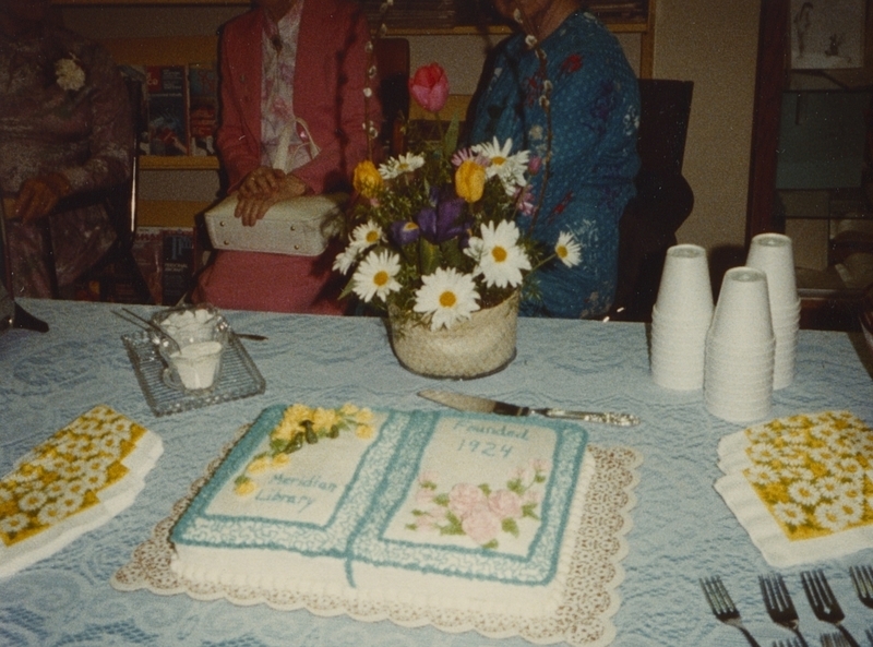 Photo of library open house cake ("Meridian Library Founded 1924") next to floral arrangement. The cake was made by Chris Mace and the back of the photo says the floral arangement created by "Nan's."