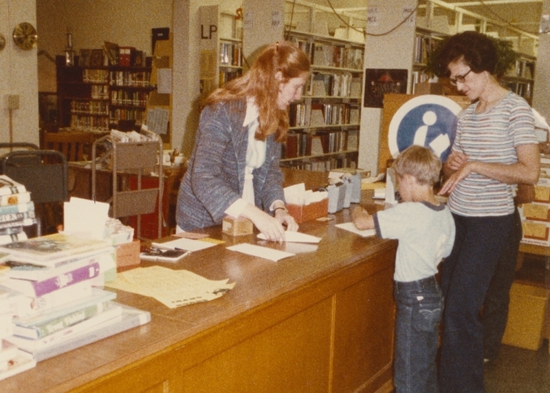 Librarian Denise works the front desk, helping a young patron. 