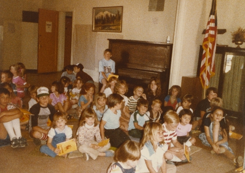 Children sit on library floor holding books, perhaps waiting for a storytime. 