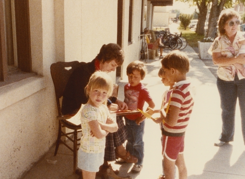 Pat, a librarian, sits outside the library with a clipboard as children gather around for an event.