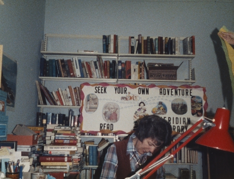 Pat Rabehl hard at work at her desk, with a "Seek Your Own Adventure--Read" parade sign in the background. 