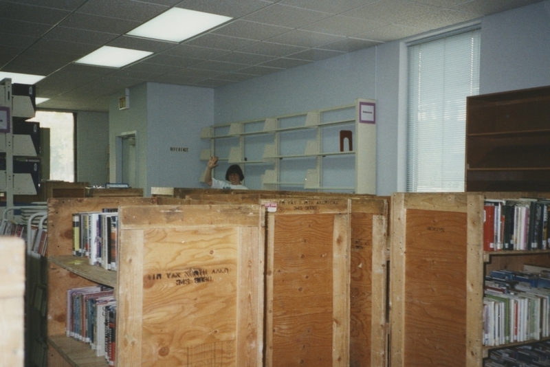 Library worker waves behind wooden crates filled with books in the adult section of the old Meridian Library. Crates prepared for move to new Cherry Lane Library building. 